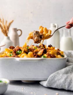 A hand lifts a serving spoon of pulled pork mac and cheese from a casserole dish.