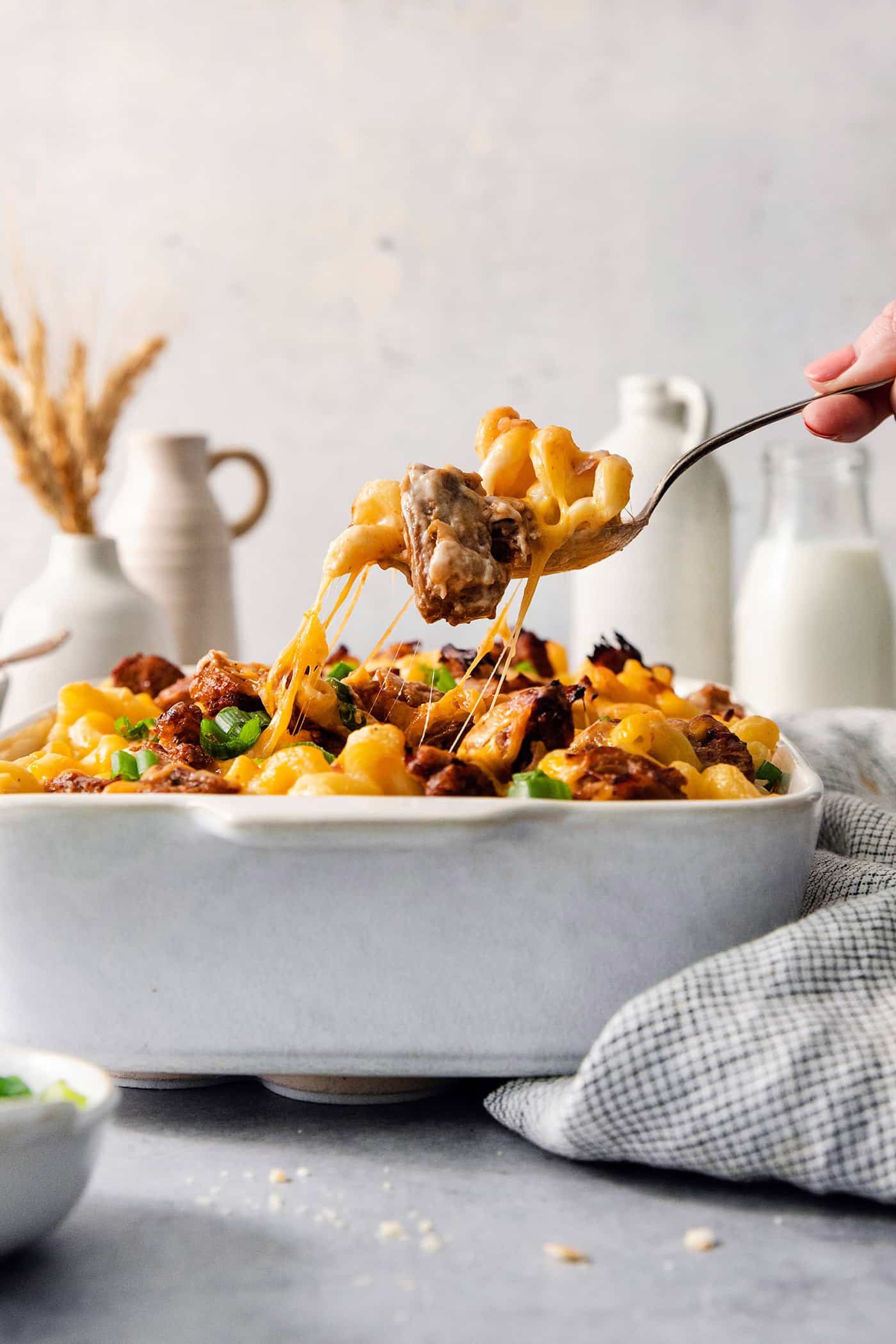 A hand lifts a serving spoon of pulled pork mac and cheese from a casserole dish.