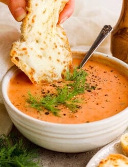 a hand dipping a piece of cheesy bread into a bowl of creamy tomato soup