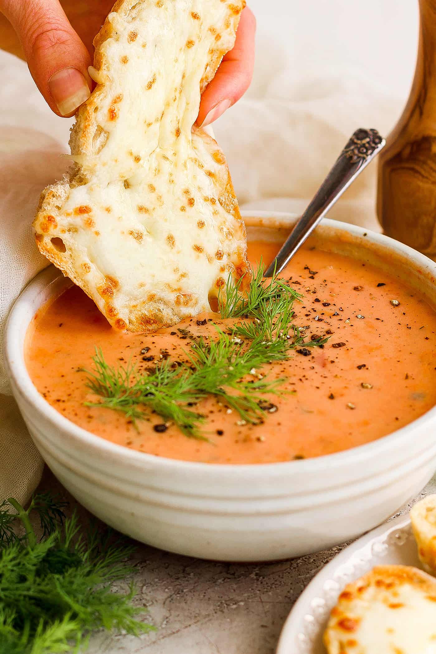 a hand dipping a piece of cheesy bread into a bowl of creamy tomato soup