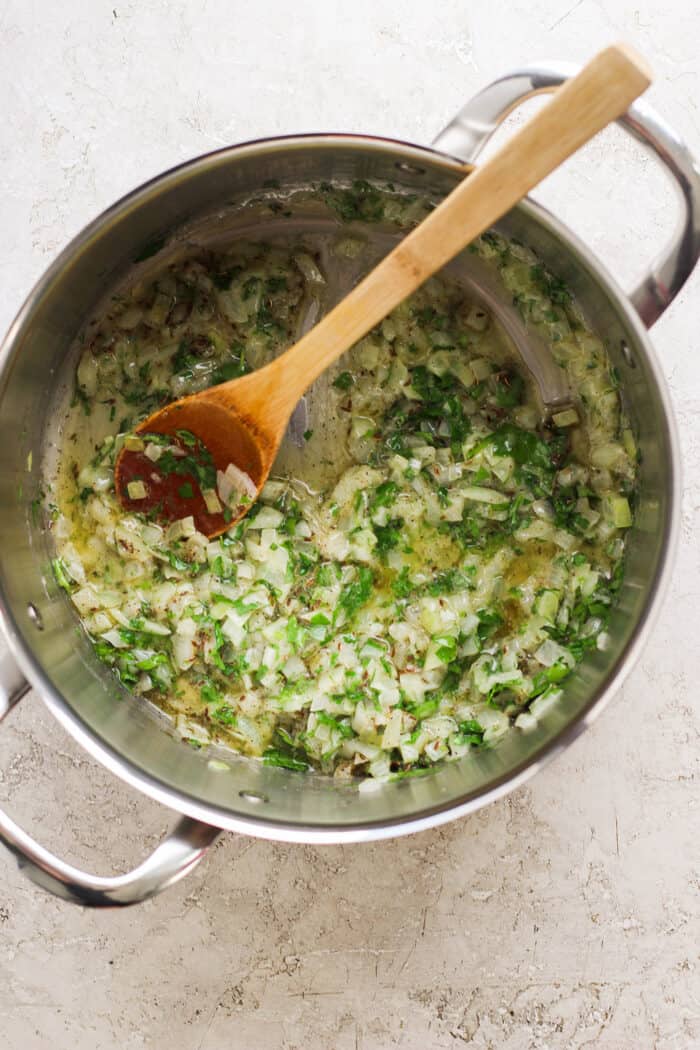 Fresh herbs, onion, and garlic are stirred with a wooden spoon in a soup pot.
