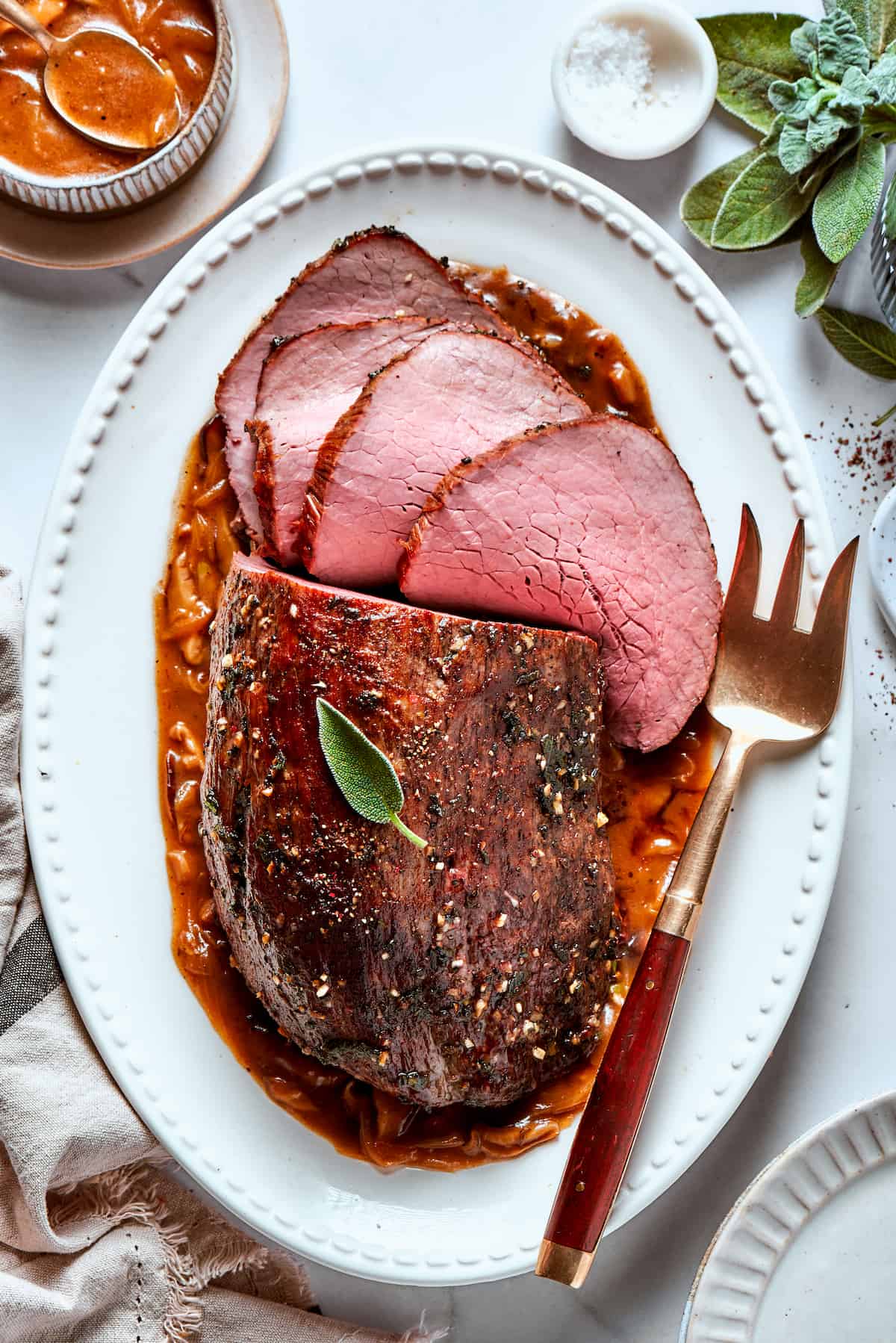 A white oval platter with a gold serving fork holds a cut top round roast.