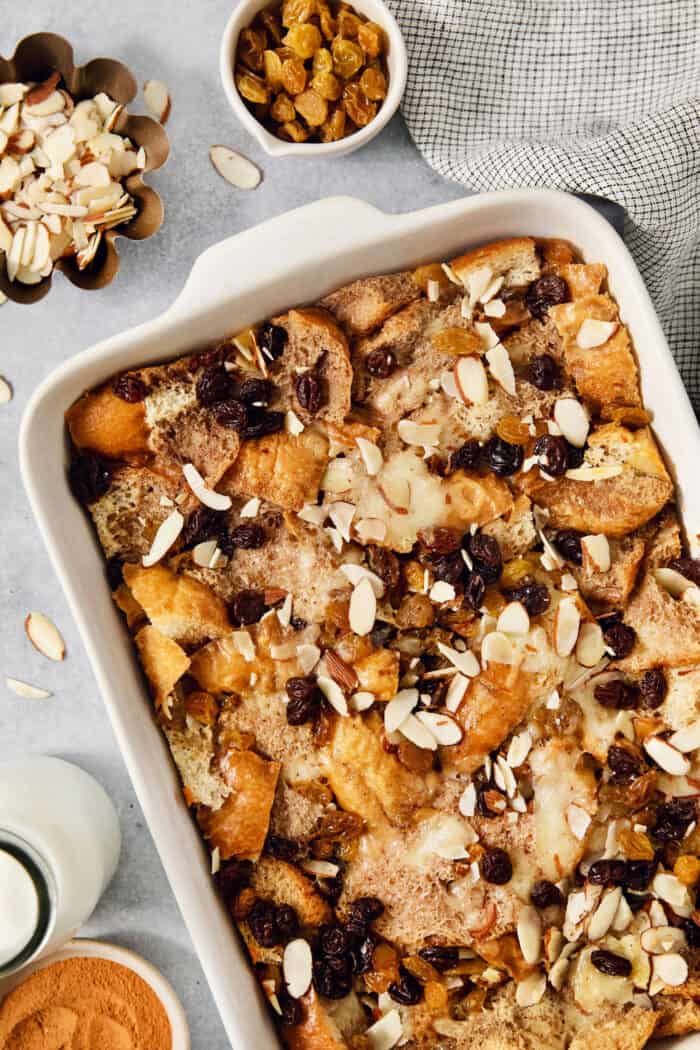 A baking dish of capirotada is surrounded by bowls of almonds and raisins.