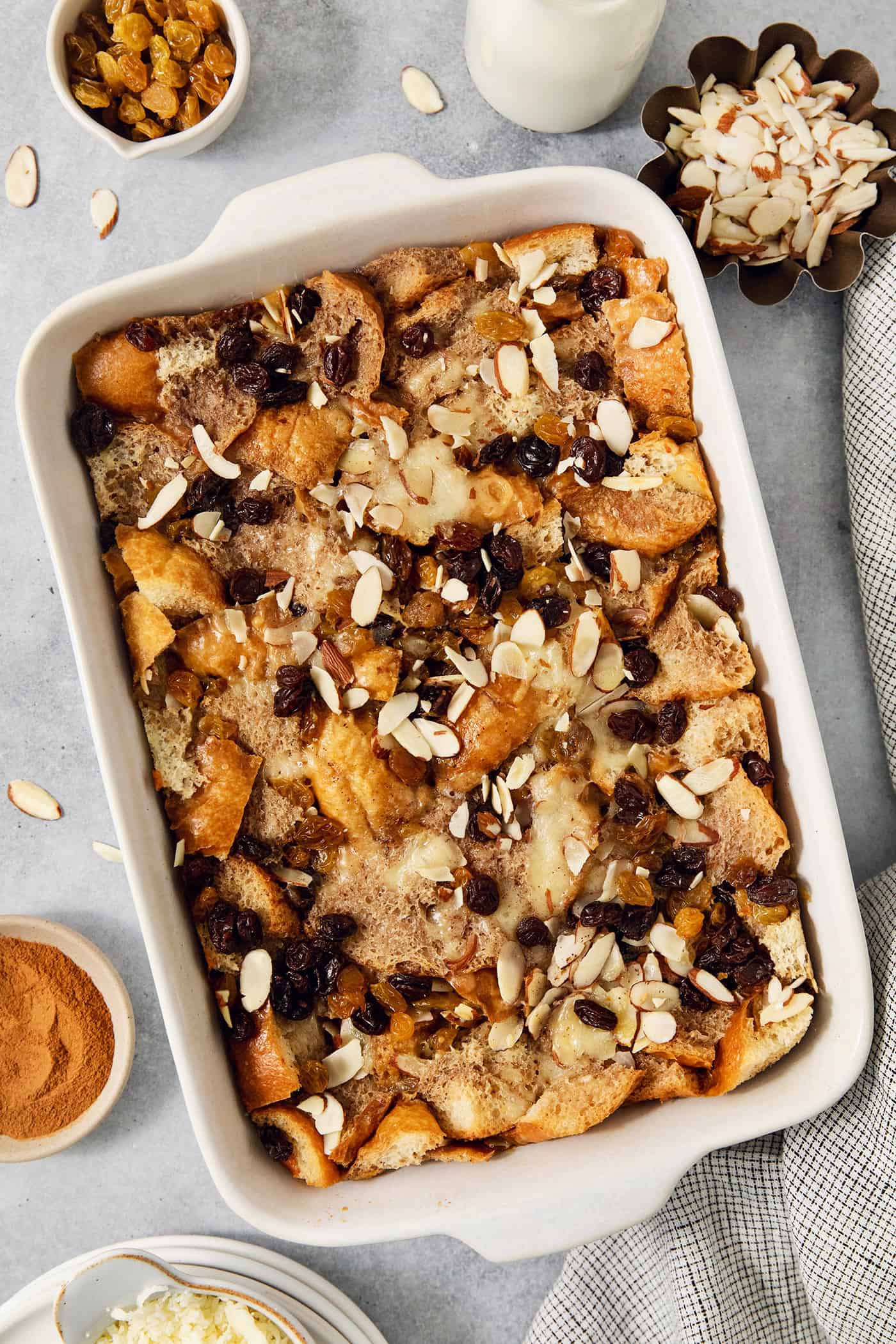 Bowls of golden raisins and almonds surround a white baking dish full of capirotada.