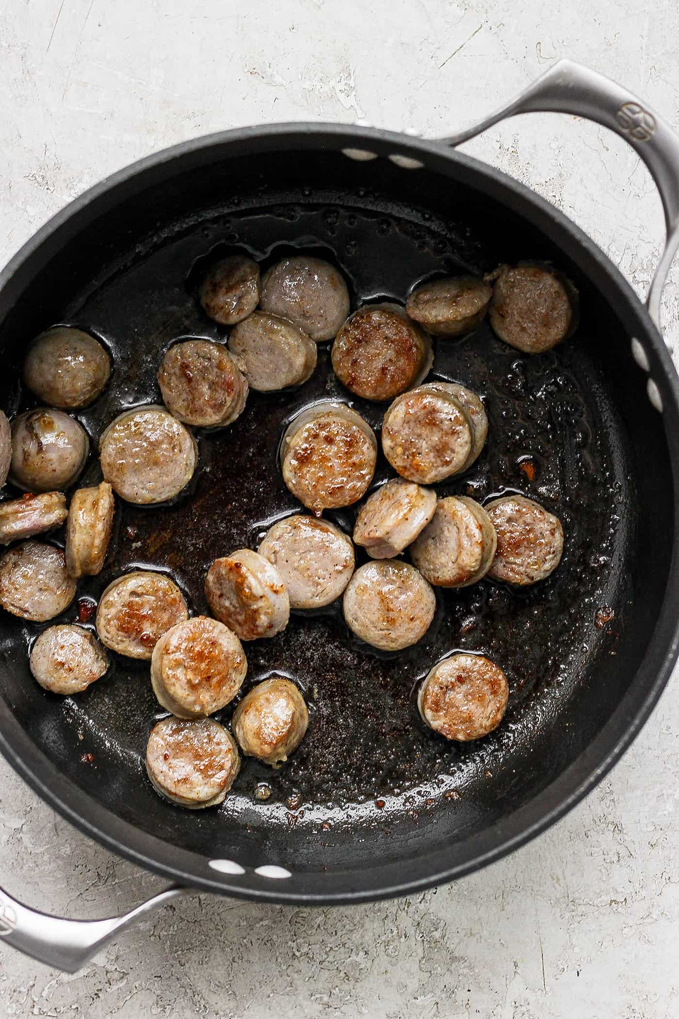 This German Potato Soup recipe is packed with savory sausage, salty bacon, and plenty of vegetables plus a spoonful of half-and-half. Slices of sausage brown in a cast iron pan.