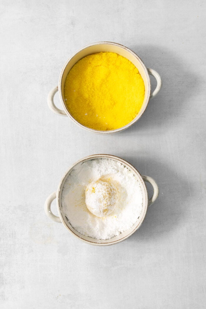 A bowl of yellow colored sugar is shown next to a bowl of powdered sugar with a cookie in it.
