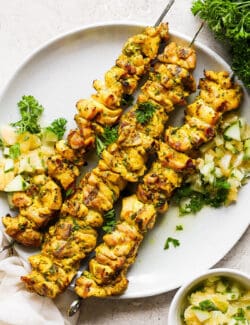 A plate holds three chicken kabobs with Meyer lemon salsa with more salsa in a bowl next to it.