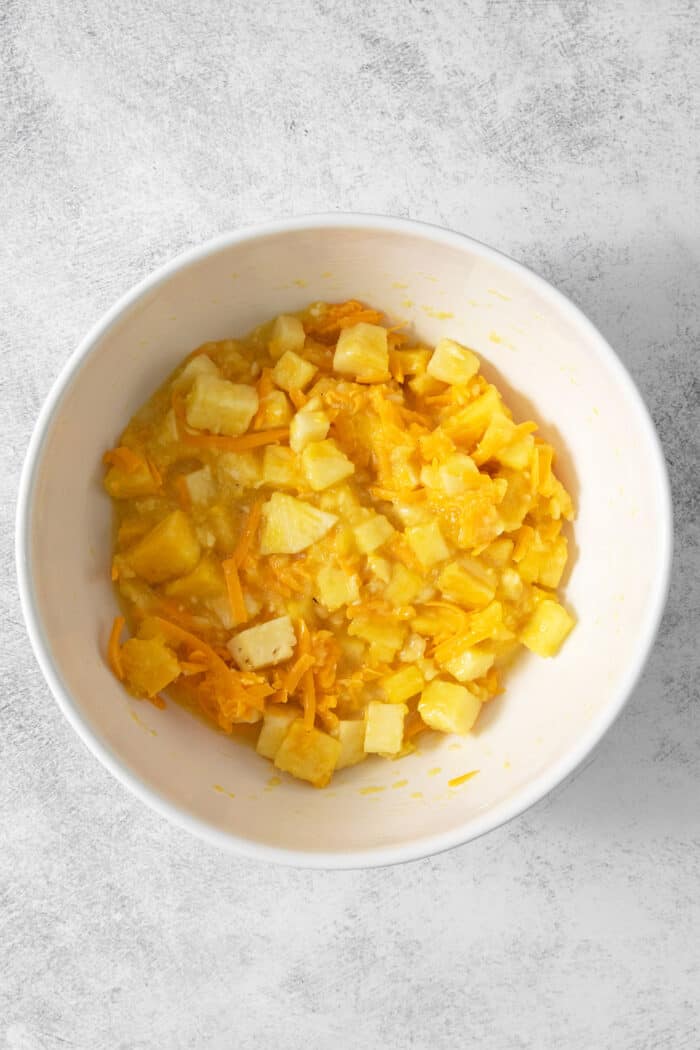 Whole pineapple chunks and shredded cheddar cheese are added to the pineapple puree in a bowl.