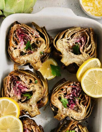 A white serving dish holds roasted artichokes with lemon slices.