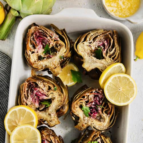 A white serving dish holds roasted artichokes with lemon slices.