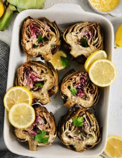 Roasted artichokes are arranged in a white serving dish and garnished with lemon slices.
