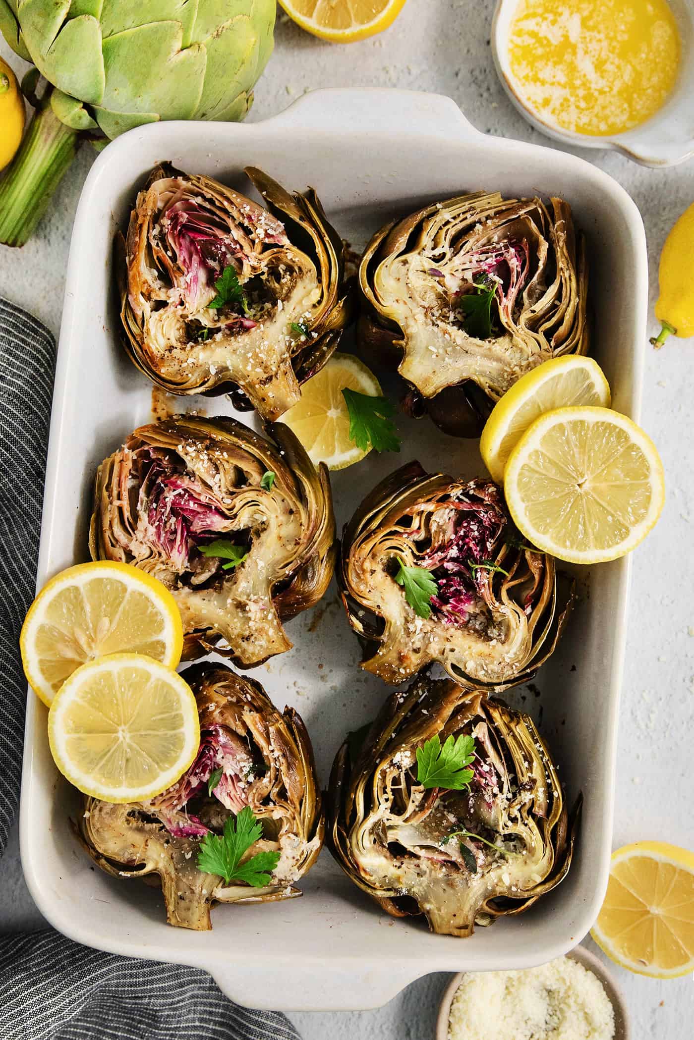 Roasted artichokes are arranged in a white serving dish and garnished with lemon slices.