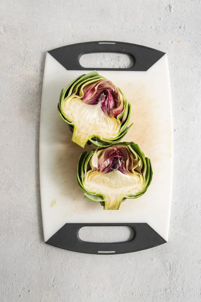 An artichoke is split in half and displayed on a white cutting board.