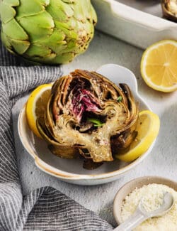 A beautifully roasted artichoke is served in a white bowl with a lemons, a raw artichoke, and seasonings around it.