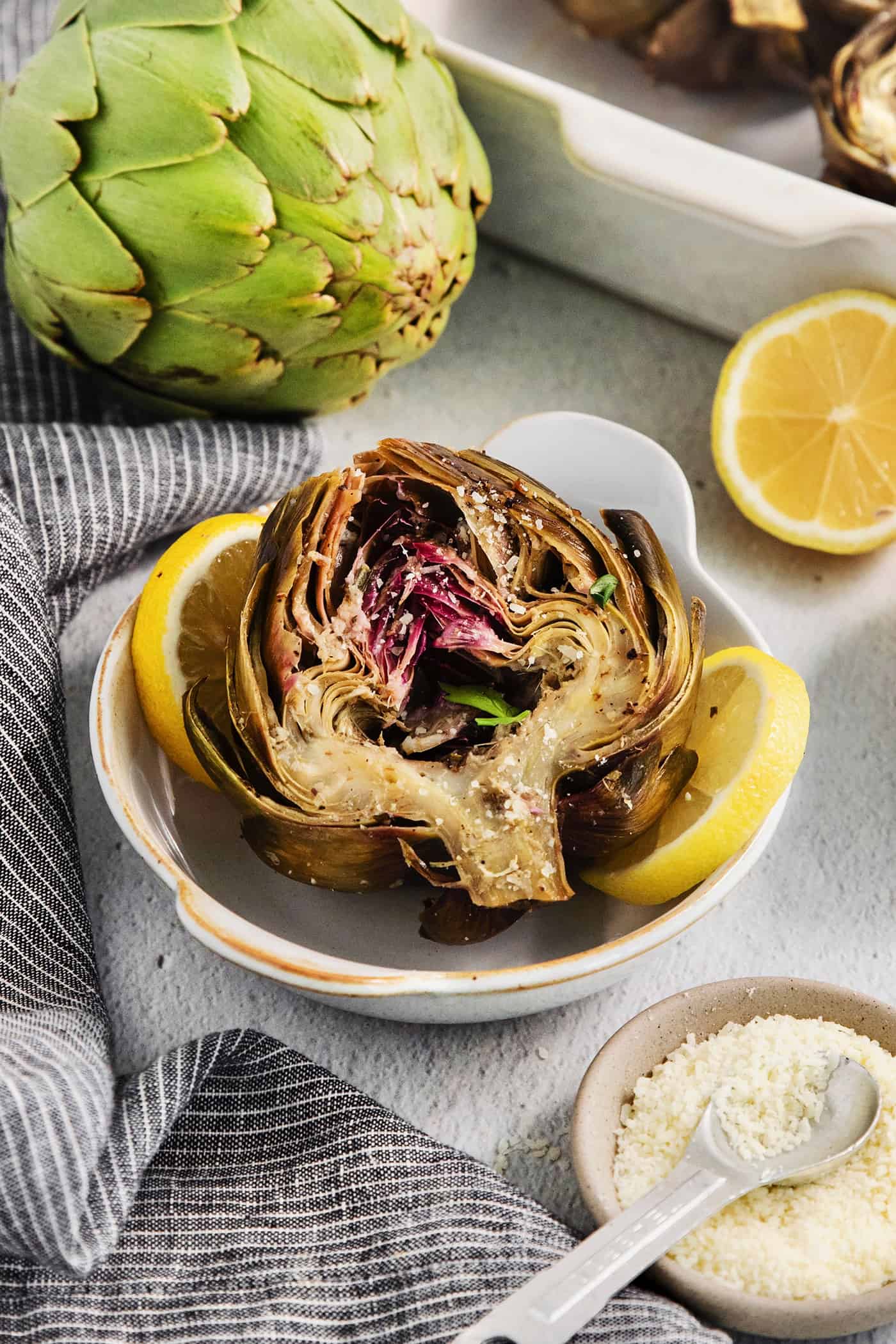 A beautifully roasted artichoke is served in a white bowl with a lemons, a raw artichoke, and seasonings around it.