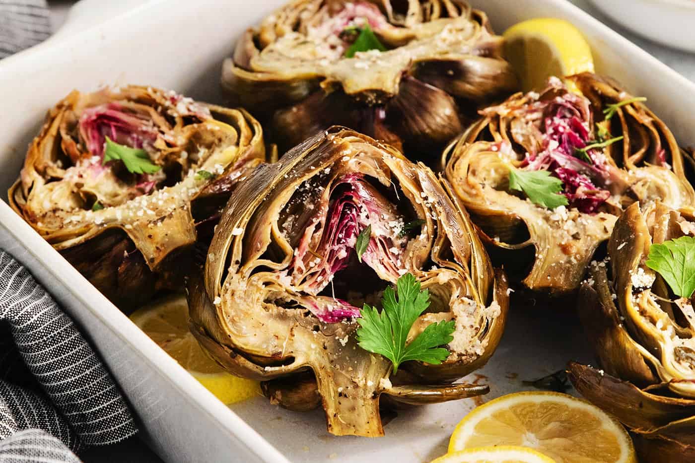 A white serving dish holds roasted artichokes with lemon slices.