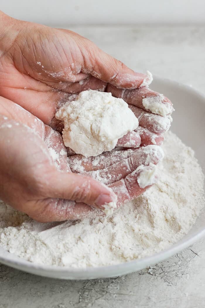 Two cupped hands hold a piece of biscuit over a bowl of flour.