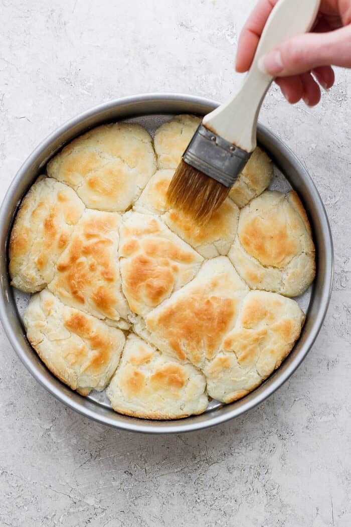 A hand uses a pastry brush to brush the baked biscuits with melted butter.