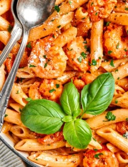 Fresh basil leaves top a serving bowl of cajun shrimp pasta with serving utensils alongside it.