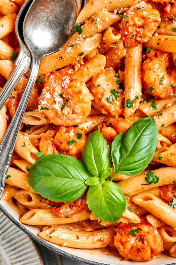 Fresh basil leaves top a serving bowl of cajun shrimp pasta with serving utensils alongside it.