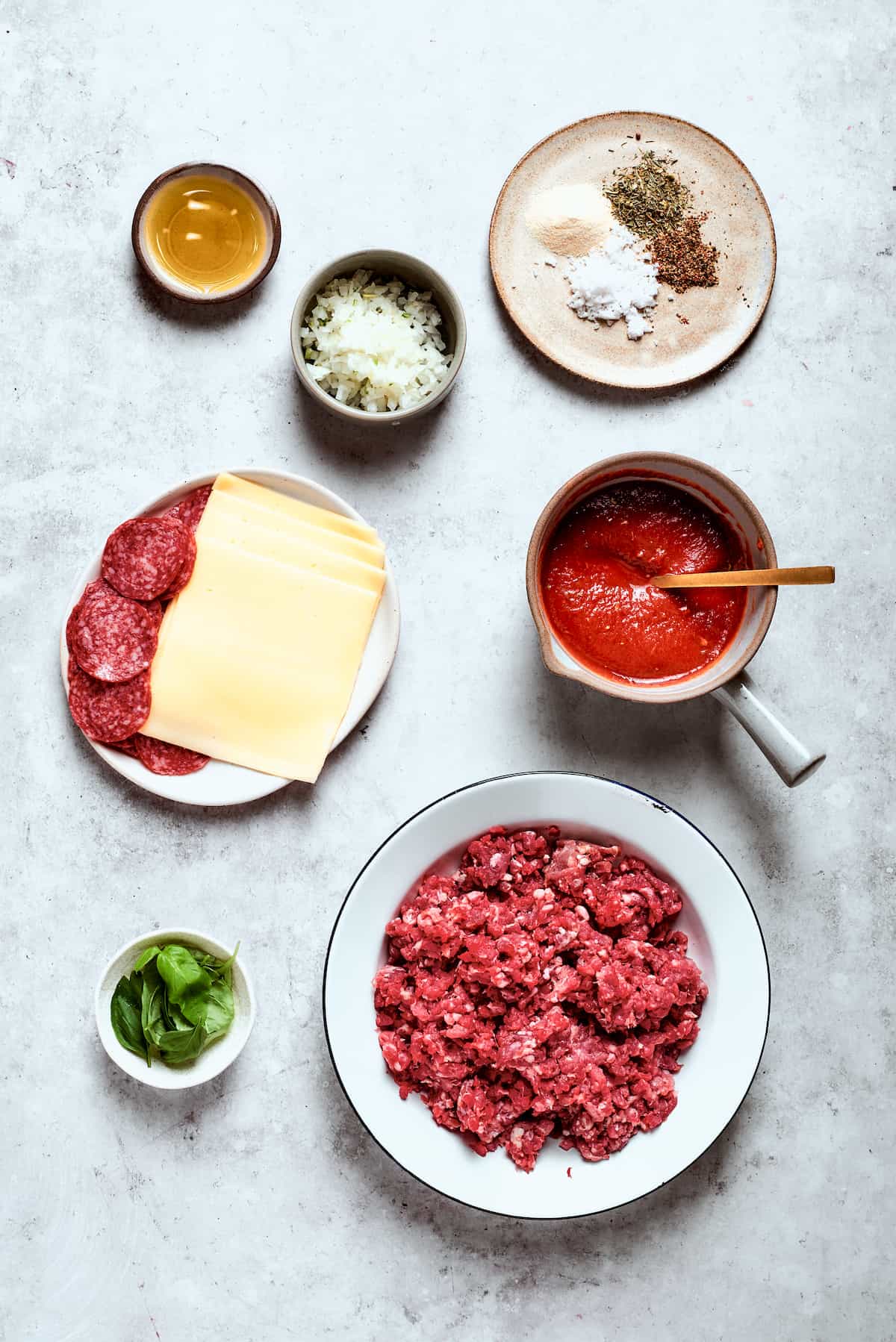 Blend two favorites to make this Pizza Burger recipe! Juicy beef patties are topped with tangy marinara, pepperoni, mozzarella & fresh basil. Ingredients for pizza burgers are shown: pepperoni, mozzarella slices, beef, tomato sauce, seasonings, basil.