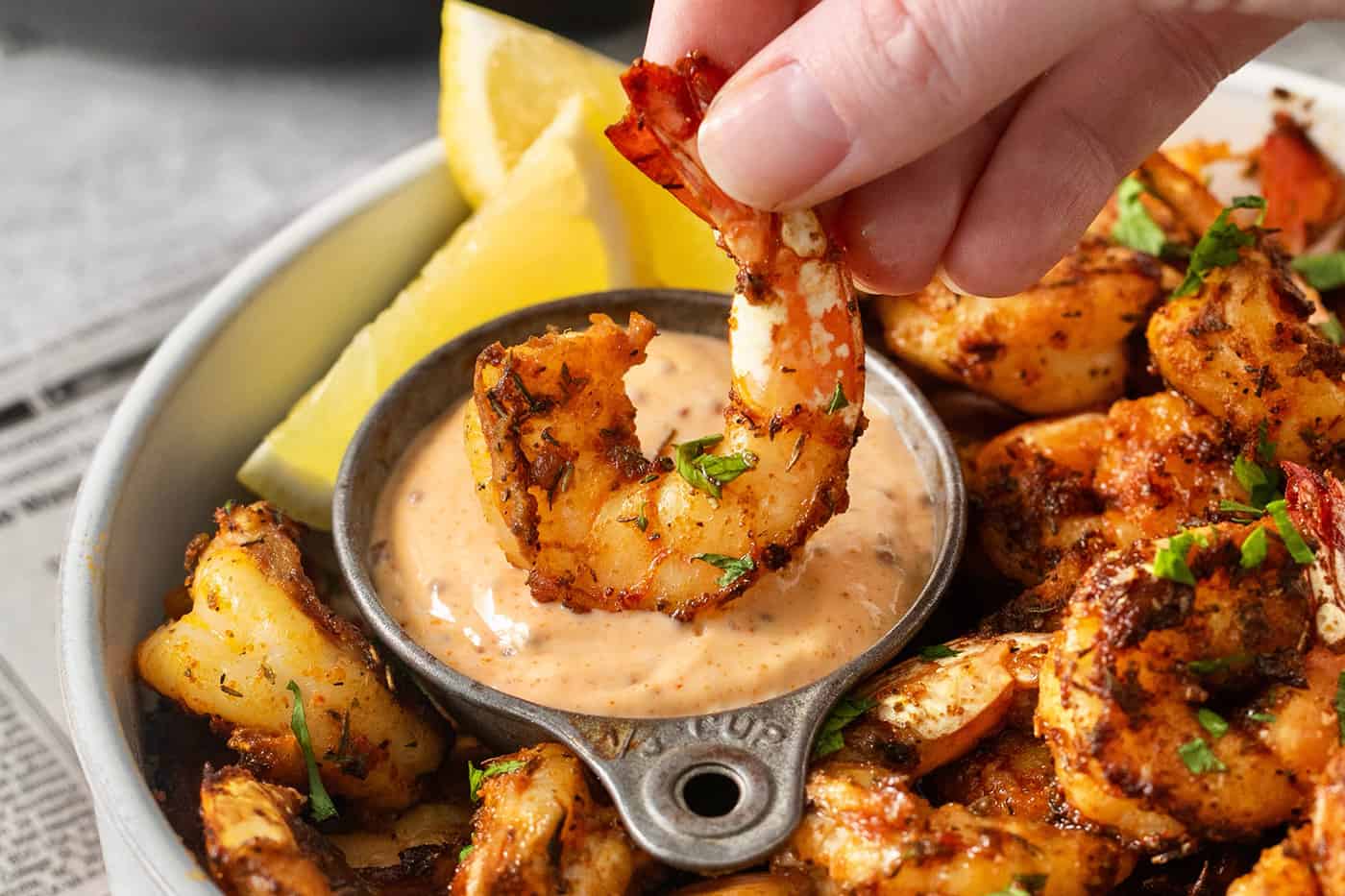 A hand dips a piece of blackened shrimp into a bowl of sauce.