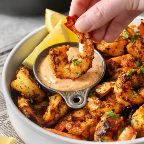 A hand dips a piece of blackened shrimp into a bowl of sauce.