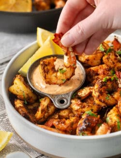 A hand dips a piece of blackened shrimp into a bowl of sauce.