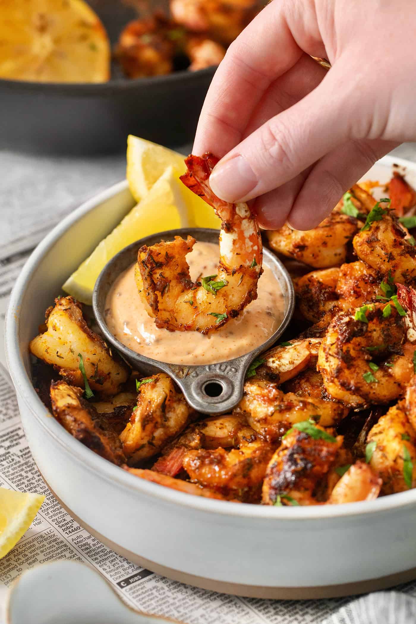 A hand dips a piece of blackened shrimp into a bowl of sauce.