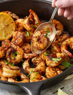 A spoon lifts out a piece of blackened shrimp from a skillet of shrimp.