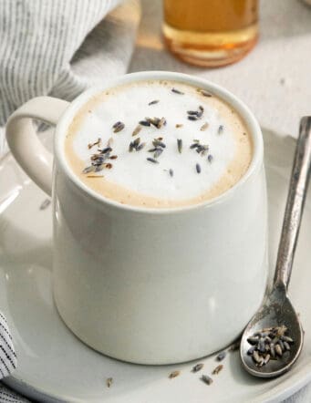 A creamy mug of lavender latte on a plate with a spoon resting next to it.