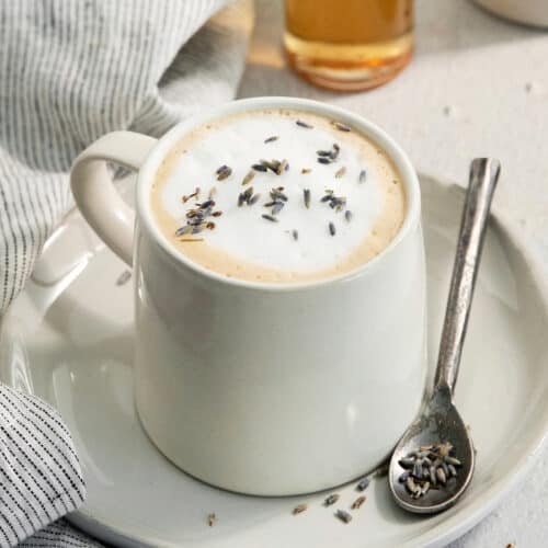 A creamy mug of lavender latte on a plate with a spoon resting next to it.