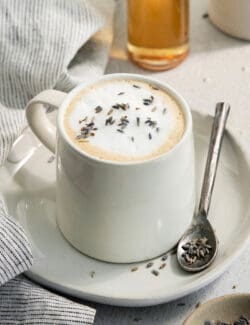 A creamy mug of lavender latte on a plate with a spoon resting next to it.