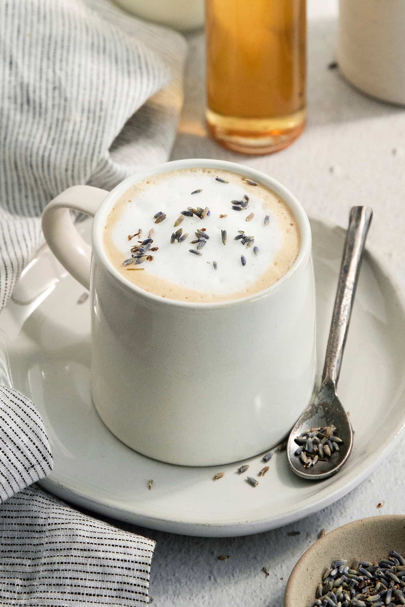 A creamy mug of lavender latte on a plate with a spoon resting next to it.