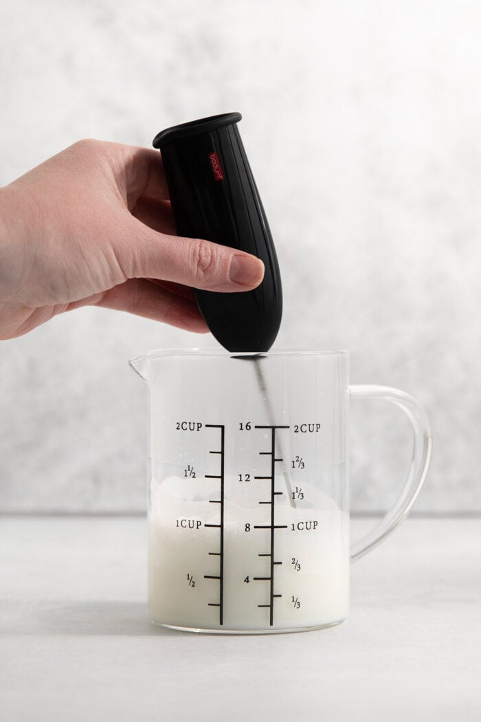 A hand holds a milk frother to froth oat milk in a glass pitcher.