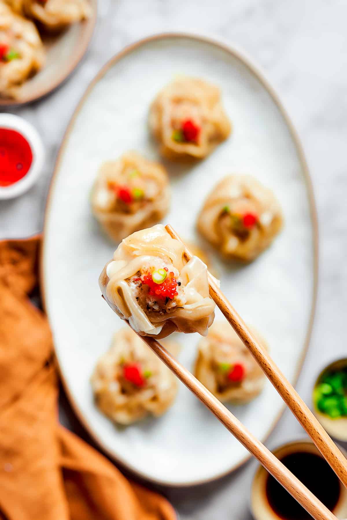 An oval serving dish holds shrimp shumai and a pair of chopsticks holds a dumpling above it.