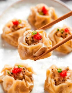 Shrimp shumai on a plate with a pair of chopsticks holding one piece above the plate.