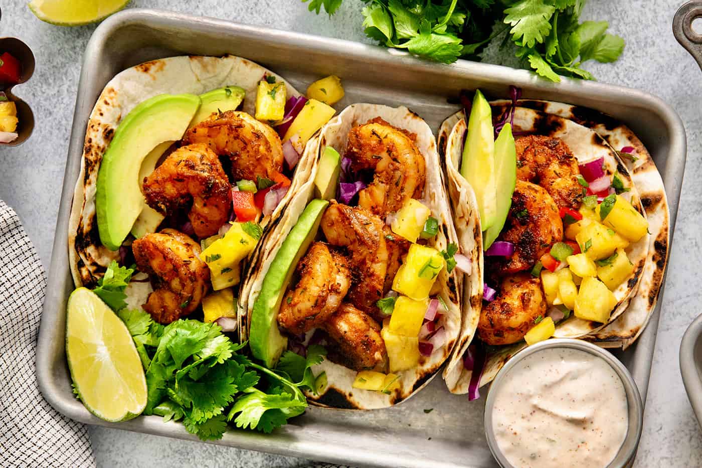 A trays holds blackened shrimp tacos surrounded by lime wedges and a bowls of avocado crema.