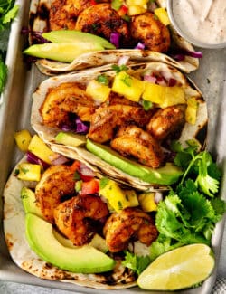 A trays holds blackened shrimp tacos surrounded by lime wedges and a bowls of avocado crema.