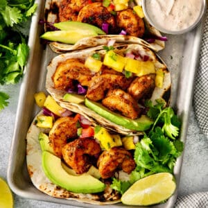 A trays holds blackened shrimp tacos surrounded by lime wedges and a bowls of avocado crema.