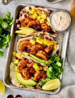 A trays holds blackened shrimp tacos surrounded by lime wedges and a bowls of avocado crema.