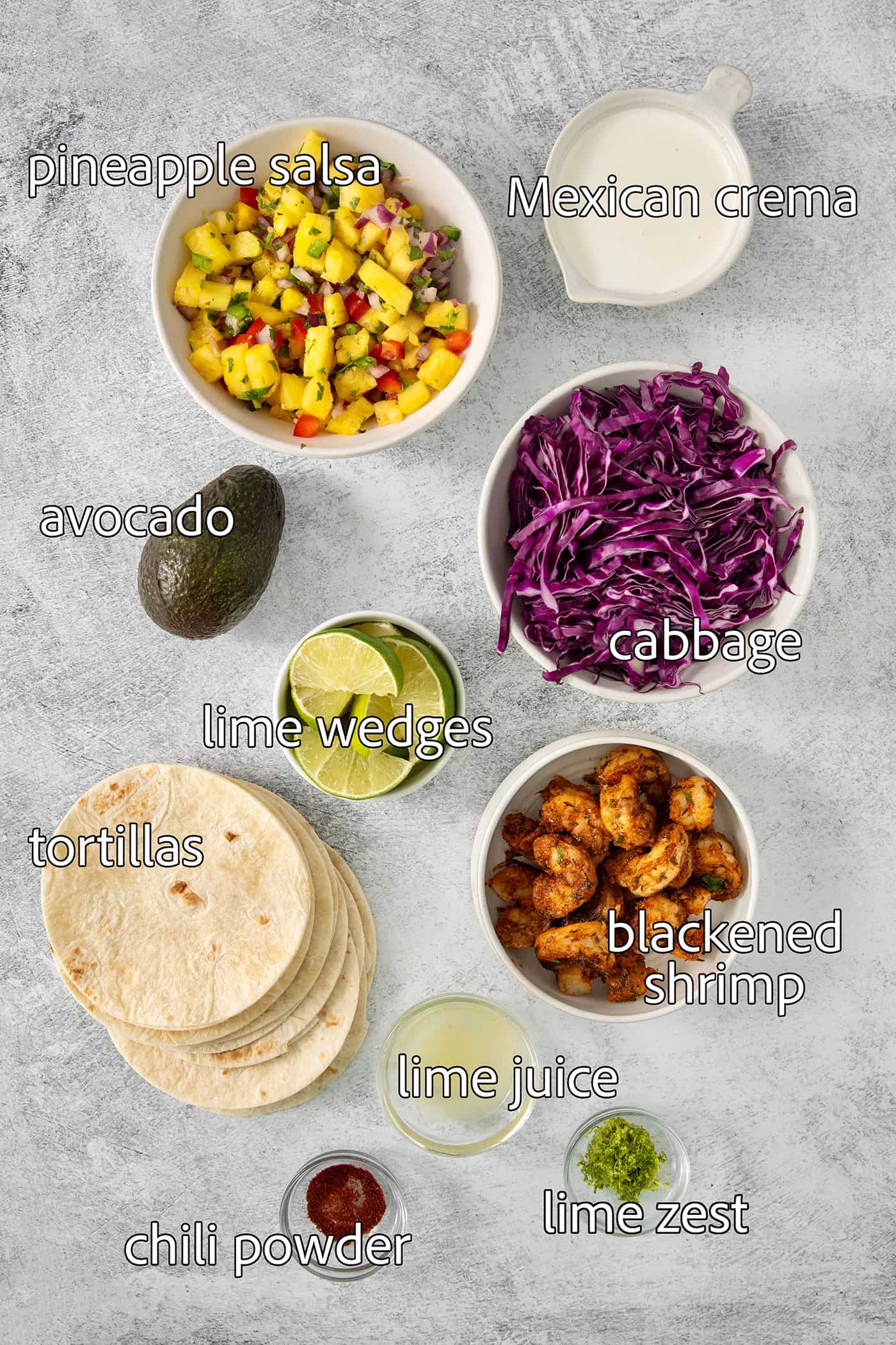 The ingredients for blackened shrimp tacos are portioned out and labeled: blackened shrimp, pineapple salsa, tortillas, cabbage, avocado, crema, lime.