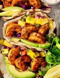 A close up shows pieces of blackened shrimp in tortillas with avocado slices and pineapple chunks.