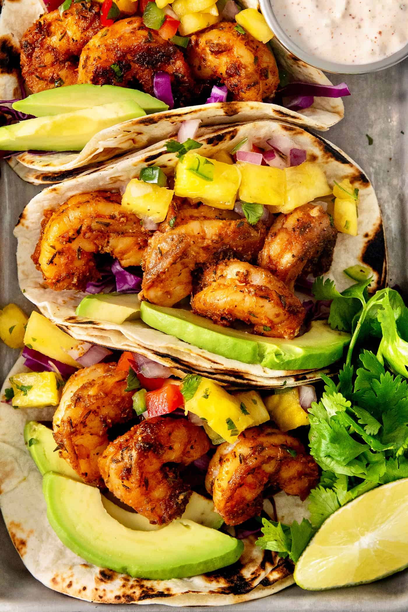A close up shows pieces of blackened shrimp in tortillas with avocado slices and pineapple chunks.