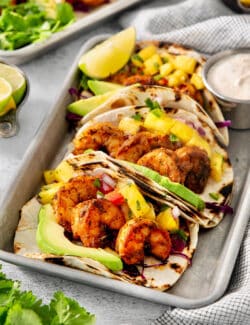 Trays of blackened shrimp tacos with lime wedges and avocado crema.
