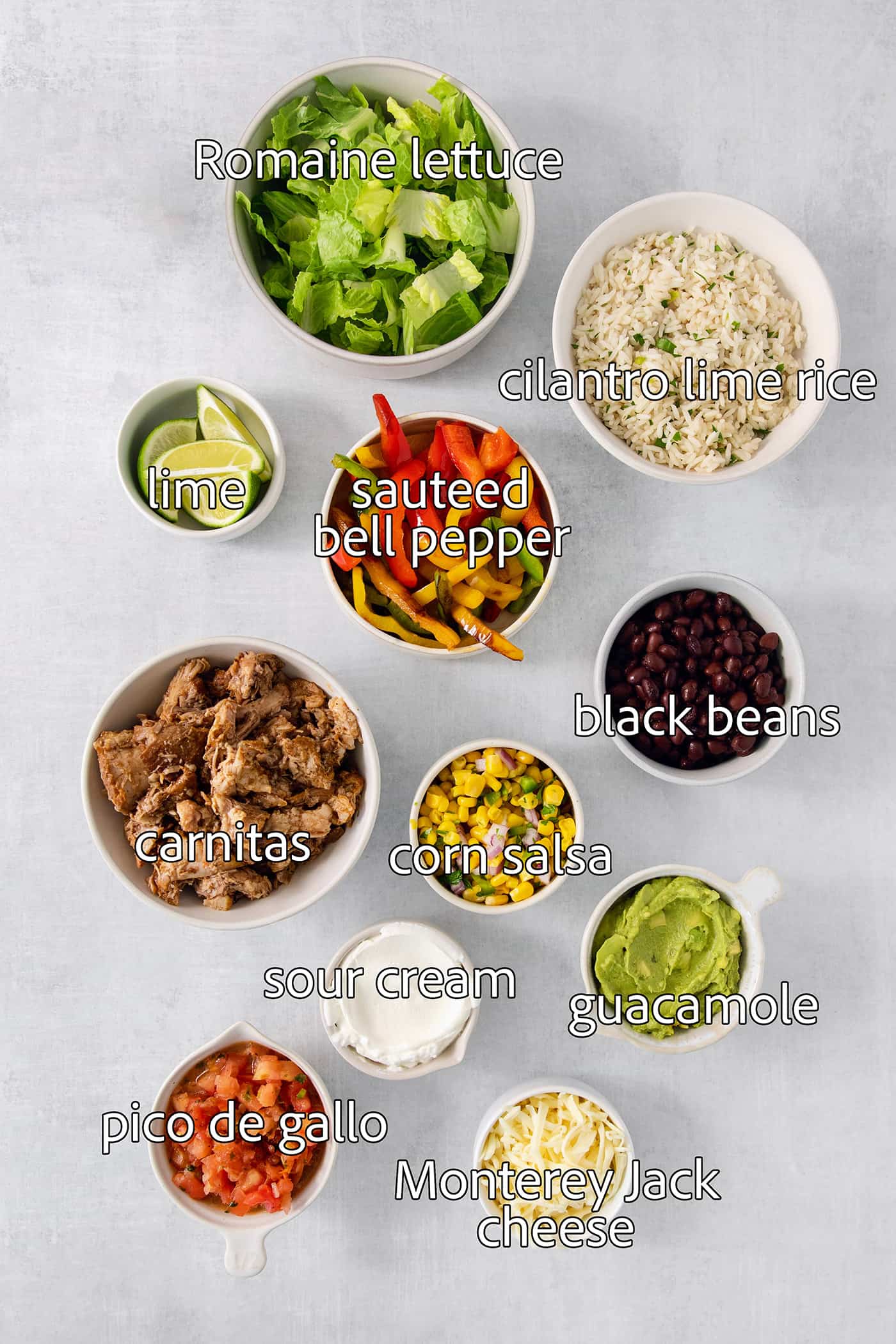 The ingredients for carnitas burrito bowls are shown portioned out and labeled: carnitas, sweet red peppers, black beans, corn salsa, guacamole, pico de gallo, lime, cilantro rice, lettuce, jack cheese, sour cream.