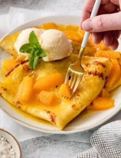 A scoop of vanilla ice cream tops crepes Suzette as a fork cuts into them.