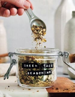A hand holding a spoon that is dropping green taco seasoning into a jar.