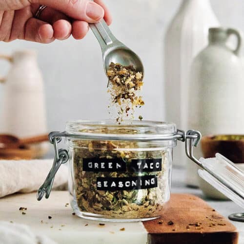 A hand holding a spoon that is dropping green taco seasoning into a jar.