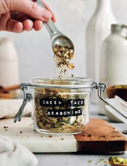 green taco seasoning in a jar, with a hand holding a measuring spoon over the top of it